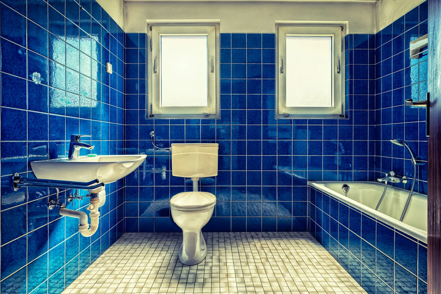 Before (real photo): a bathroom tiled floor to ceiling in glossy cobalt-blue tiles with a cream mosaic floor, a wall-hung basin with exposed pipes, a toilet with an exposed cistern between two small windows, and a tiled-in bath along the right wall