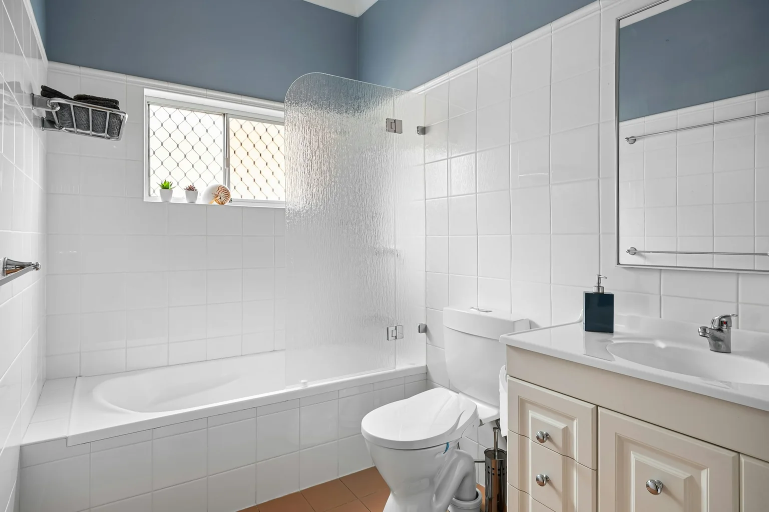 Before (real photo): a white-tiled 1990s bathroom with blue-grey paint above, a tiled bath hob with a rippled glass bath screen, a window with a diamond security grille, a cream vanity with raised-panel doors and a terracotta floor