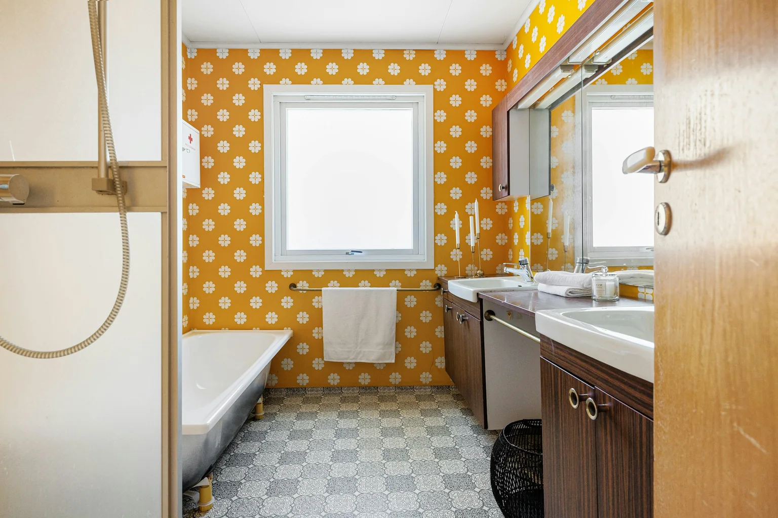 Before (real photo): a long bathroom with mustard flower-pattern walls, a bath along the left wall, a brown woodgrain double vanity with ring pulls, a mirror cabinet with a strip light and a patterned vinyl floor, seen past a shower screen and an open door