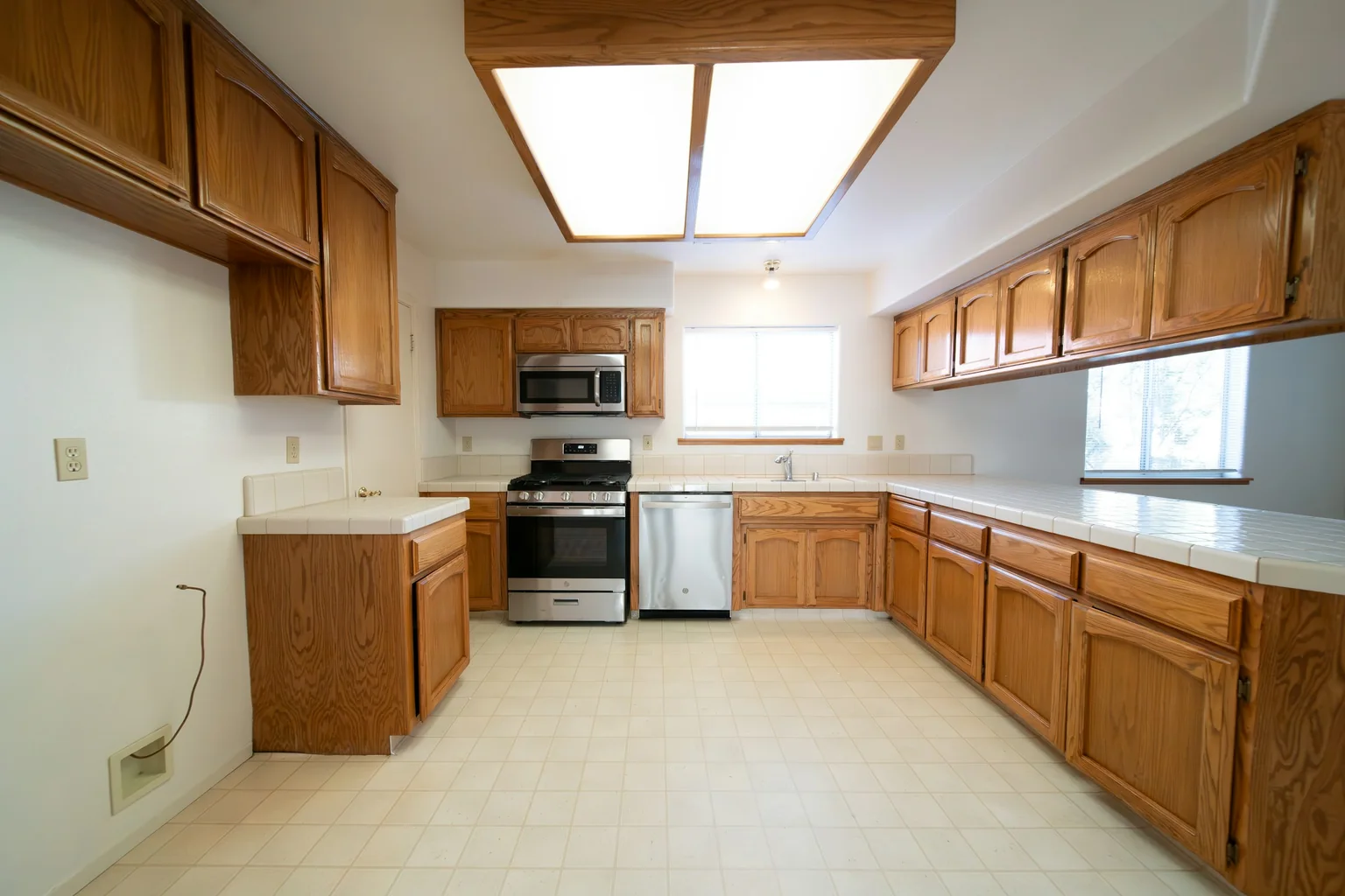 Before (real photo): a U-shaped kitchen with honey oak cathedral-arch doors, white tiled benchtops, a timber-framed fluorescent light box, a microwave over a stainless cooker and a beige vinyl tile floor