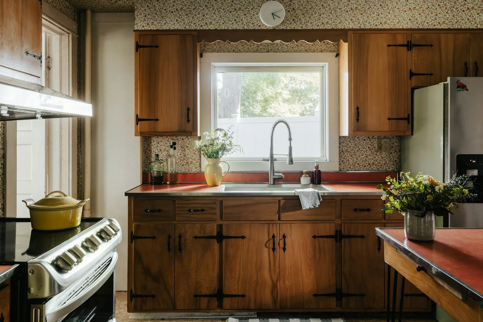 Before (real photo): a cottage kitchen with timber cupboards and black strap hinges, a red laminate benchtop with a metal edge, floral wallpaper and a scalloped pelmet over the window, a stainless cooker on the left, a fridge on the right and a small red-topped table
