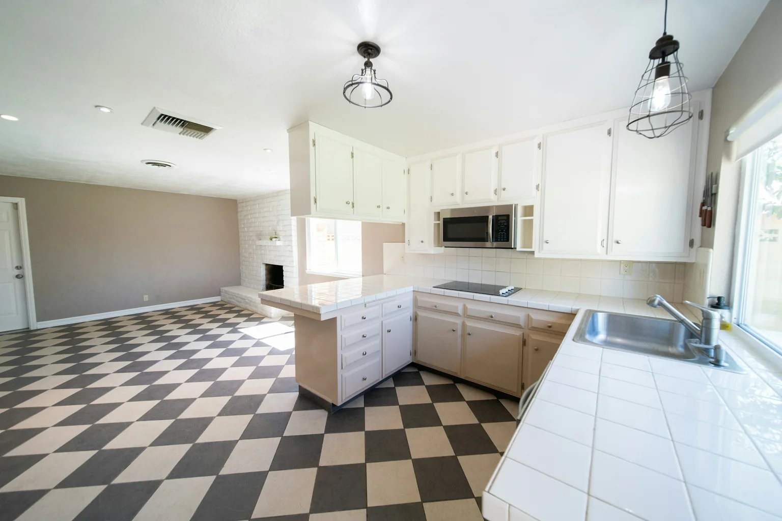 Before (real photo): an open-plan kitchen with white-painted cupboards, white tiled benchtops and a tiled raised bar on the peninsula, two cage pendants, a black and white chequerboard floor and a painted brick fireplace in the family room