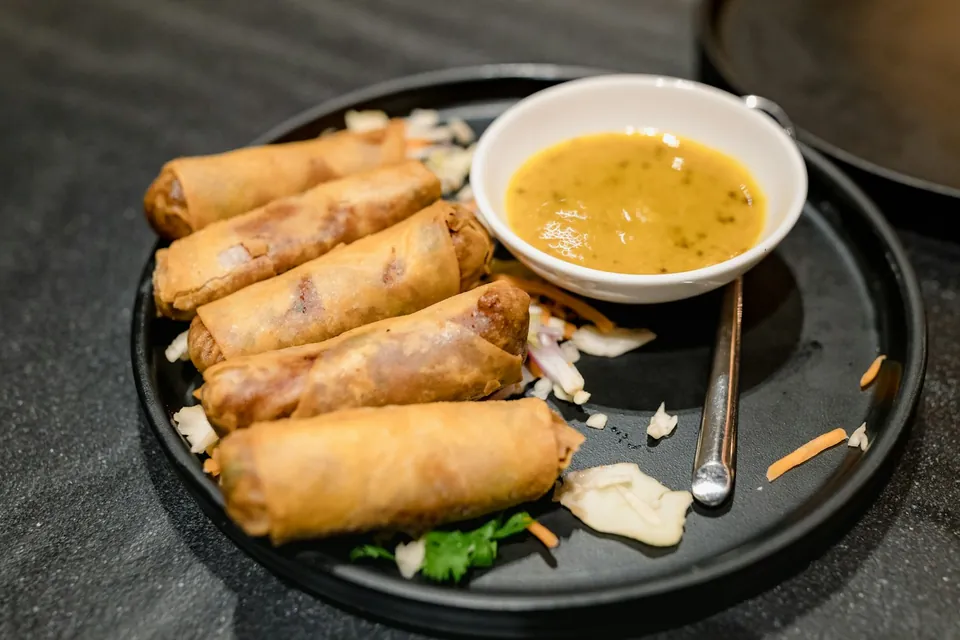 Five fried spring rolls on a dark plate with a bowl of dipping sauce