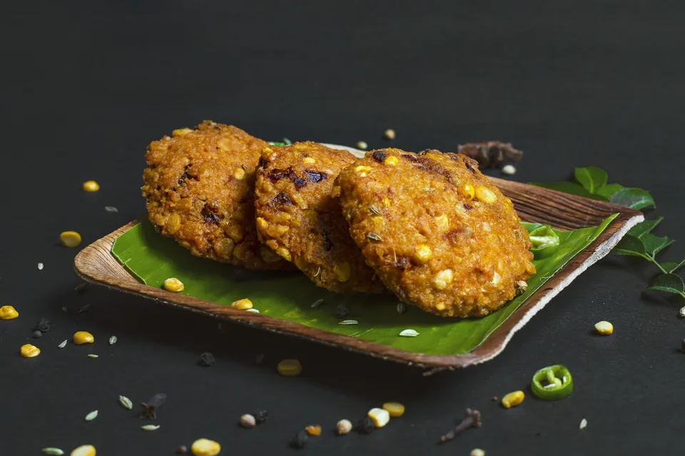 Three golden corn fritters on a banana leaf, with corn kernels and green chilli scattered around