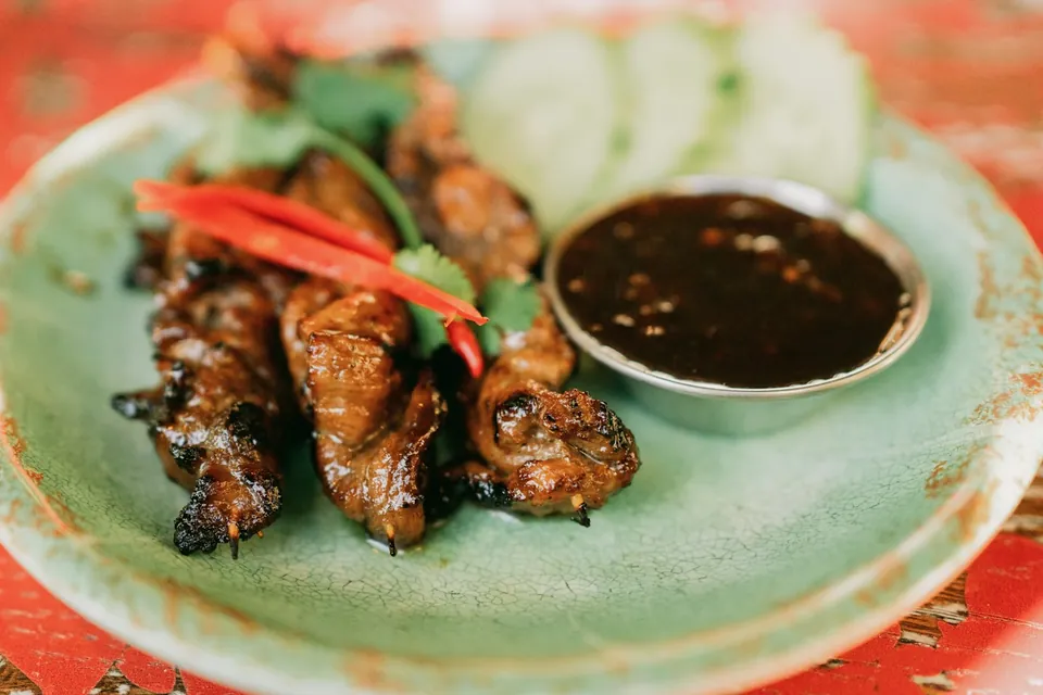 Charcoal-grilled pork skewers with a dish of dark jaew dipping sauce, cucumber and red chilli on a green plate