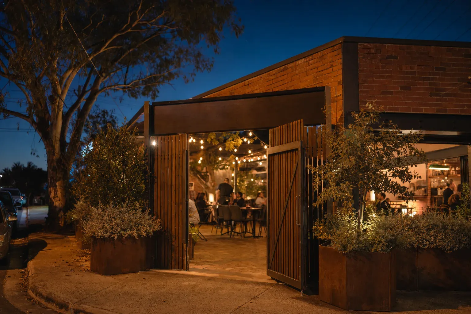 The restaurant frontage at dusk: a brick corner building with its timber gate open onto a festoon-lit courtyard