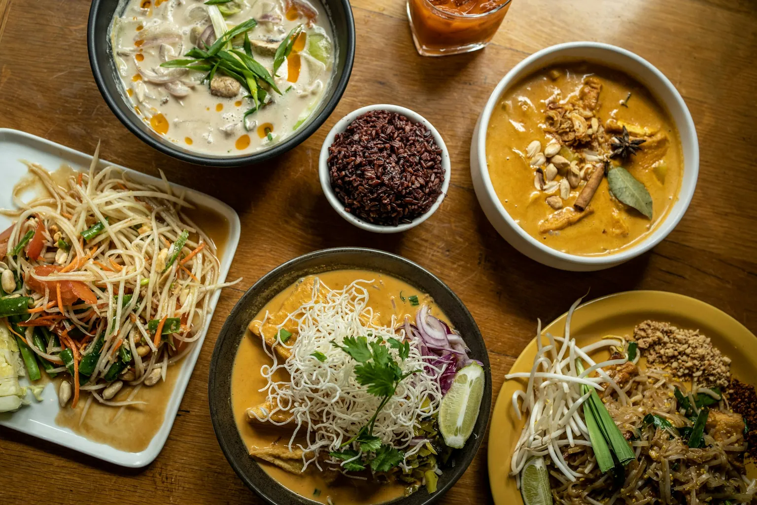A shared table from above: tom kha, a bowl of riceberry rice, massaman, som tam, khao soi and pad thai on a timber table
